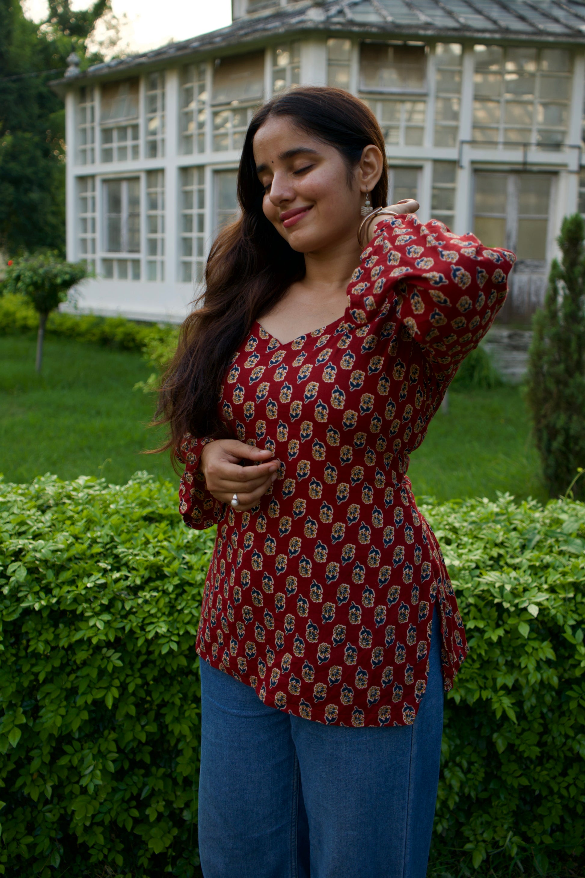 Woman in a red patterned top and blue pants standing in a garden.